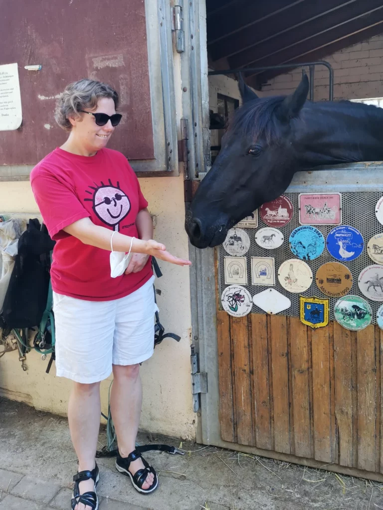 Eine Frau mit Behinderung hält ihre Hand flach einem Pferd entgegen, das neugierig aus der Stallbox schaut.